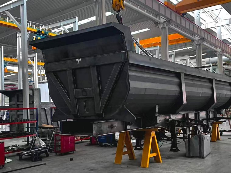 A large fabricated steel body resting on stands inside a workshop, with lifting equipment overhead.