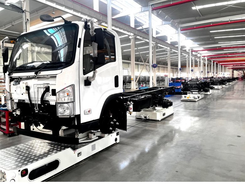 A white light-duty truck cab and chassis on an assembly line inside a plant, with further chassis units queued behind it.